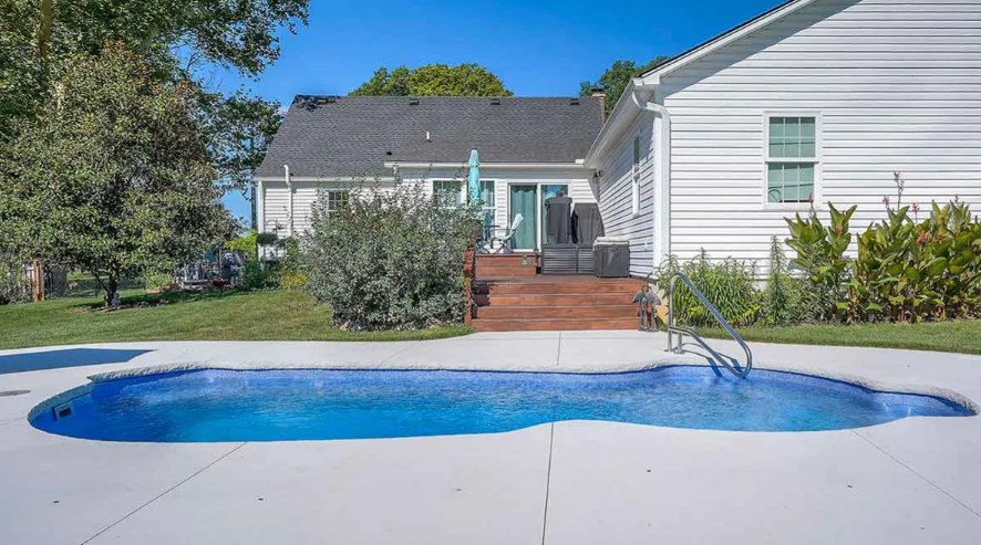Backyard with a blue pool, deck, and white house on a sunny day.
