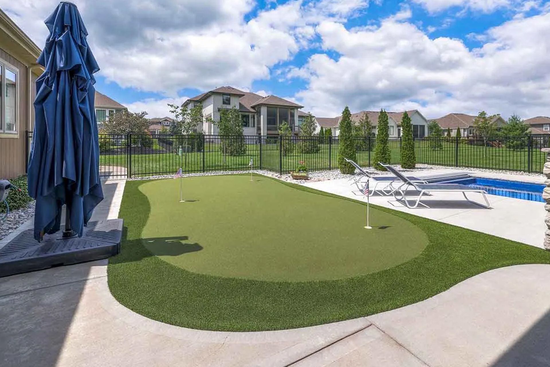 Backyard with putting green, pool, and patio. Blue umbrella, black fence, and sunny sky.