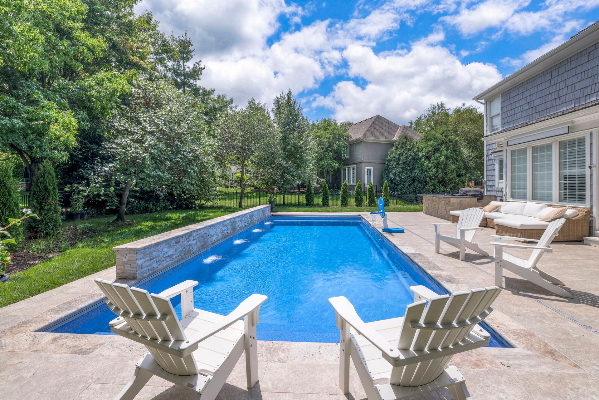 Backyard pool with white chairs, lush trees, and a house under a bright, cloudy sky.