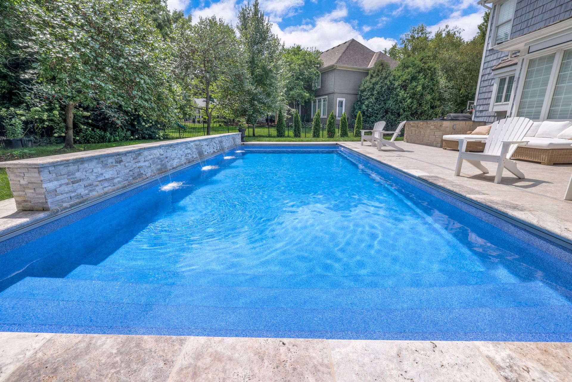 Blue rectangular swimming pool with stone deck, surrounded by trees and a house.