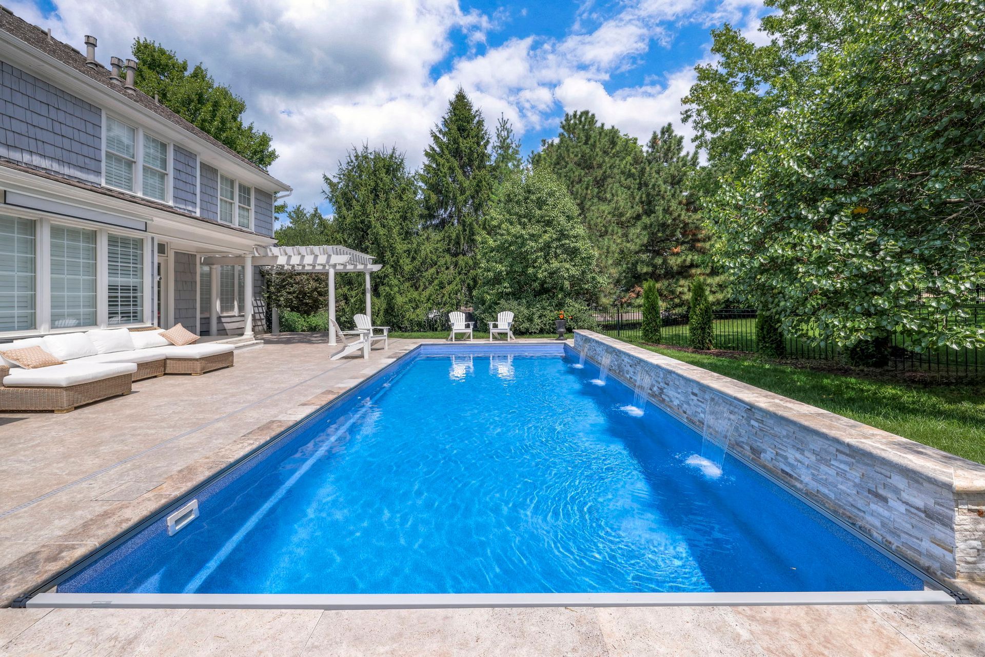 Rectangular blue swimming pool beside a gray stone patio and two-story house, with trees in the background.