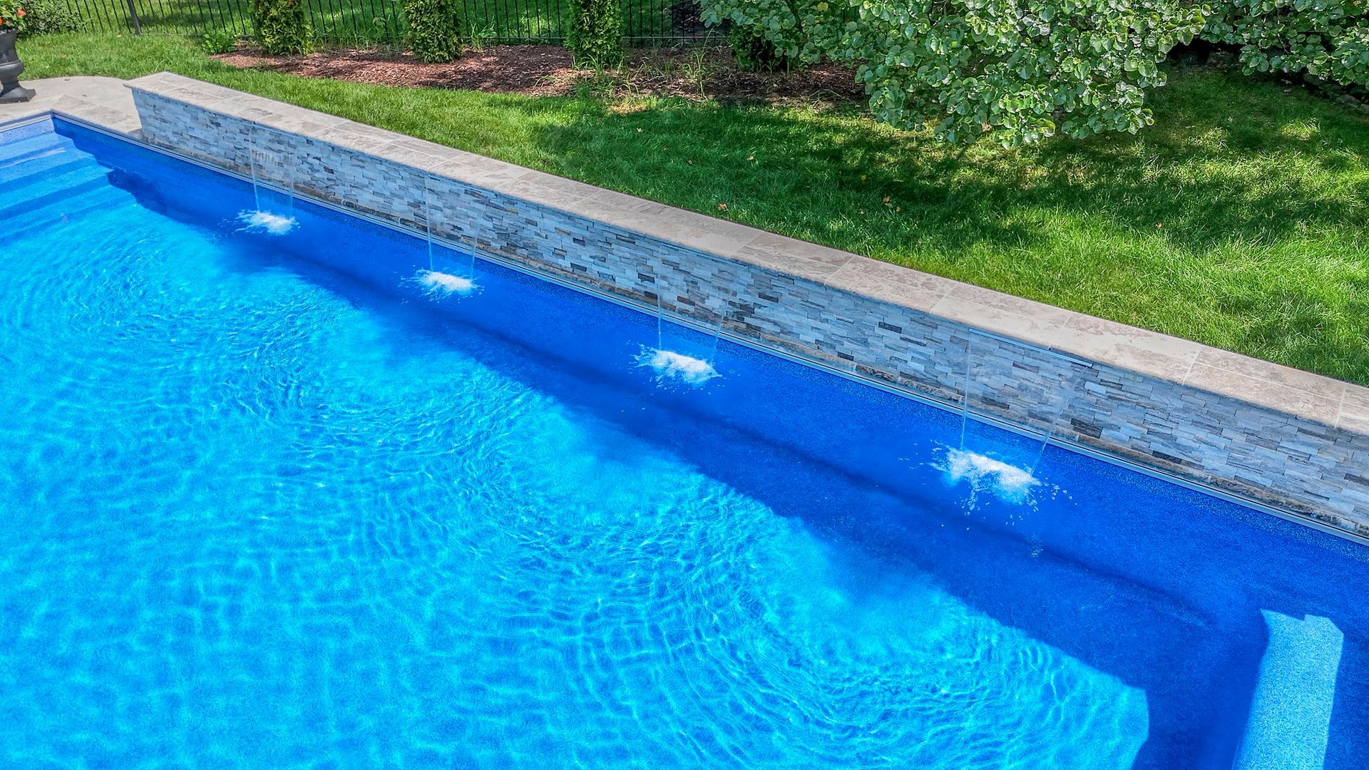 Blue pool with water jets along a textured concrete wall, surrounded by green grass and trees.