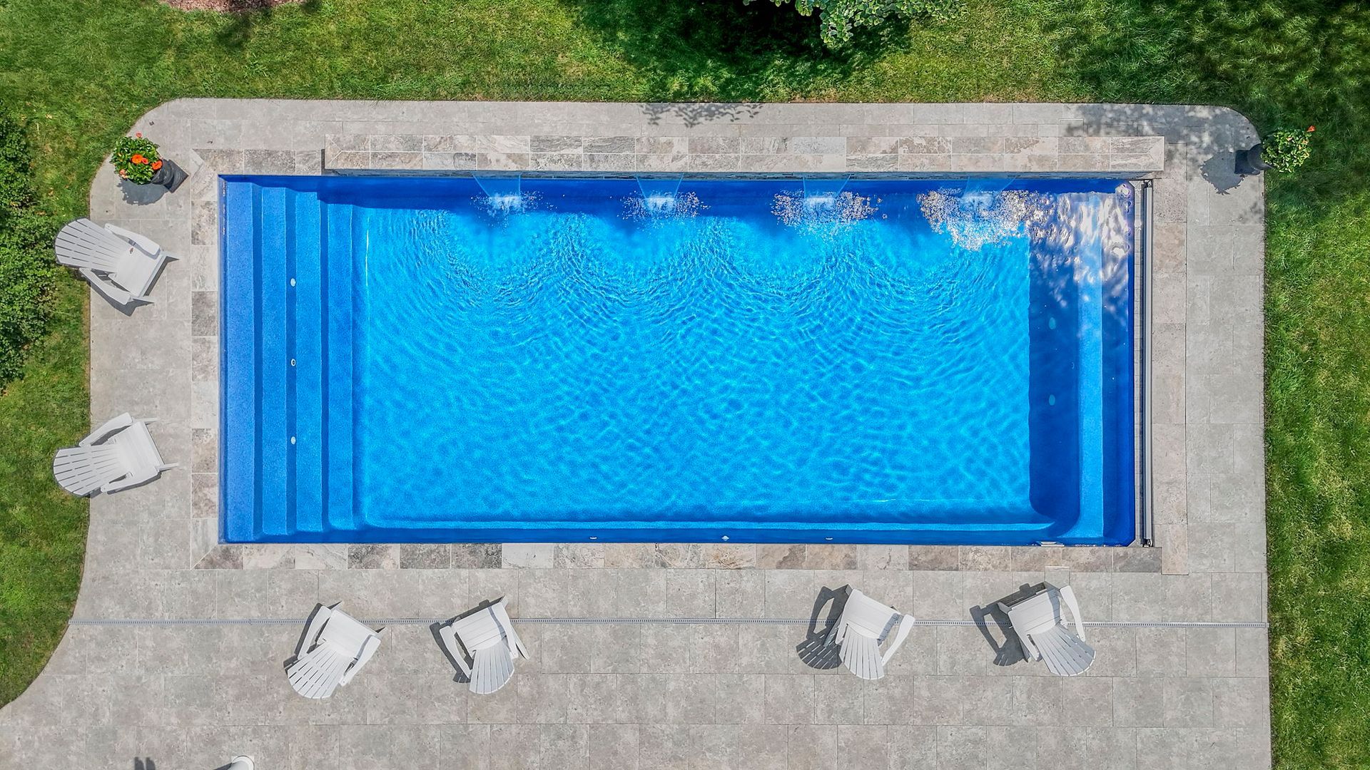 Rectangular blue swimming pool with steps, surrounded by stone patio, lawn, and lounge chairs.