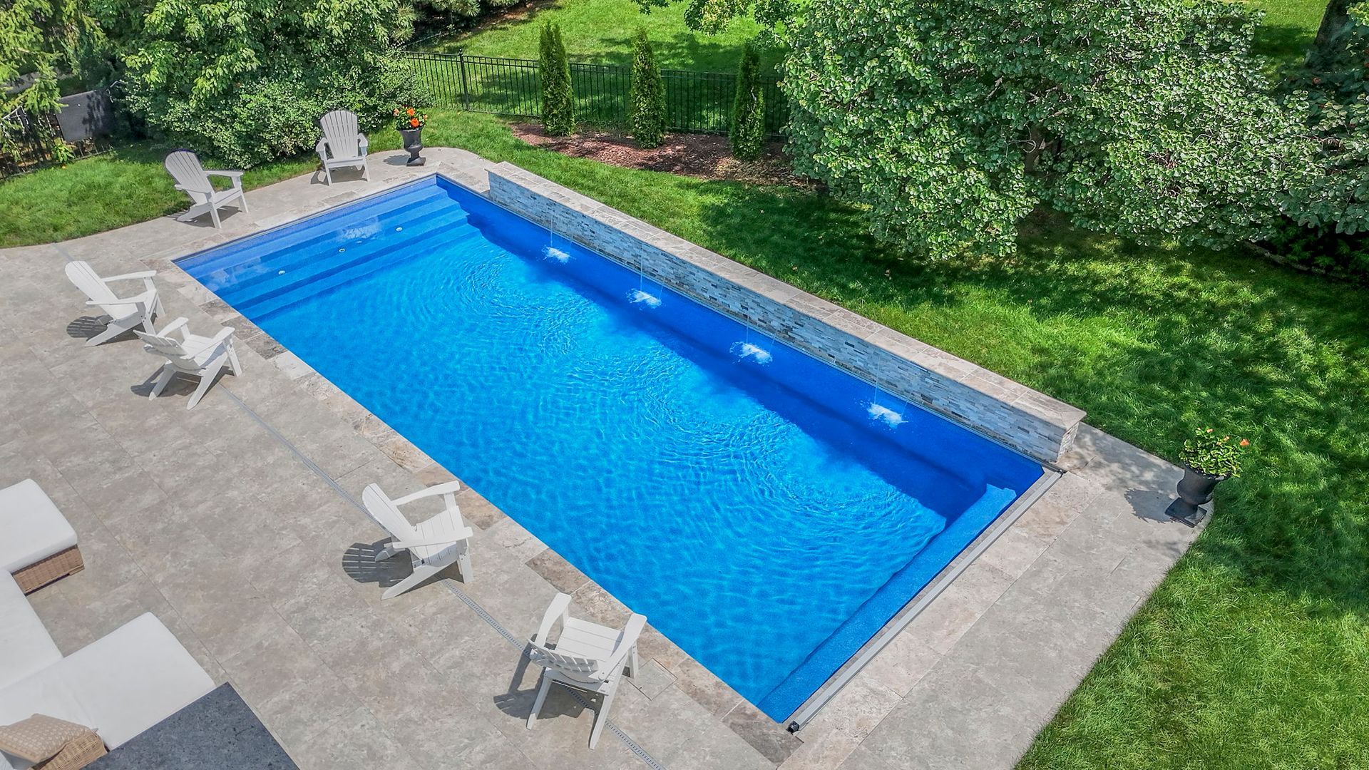 Rectangular in-ground swimming pool with blue water, concrete patio, white chairs, and grassy yard.