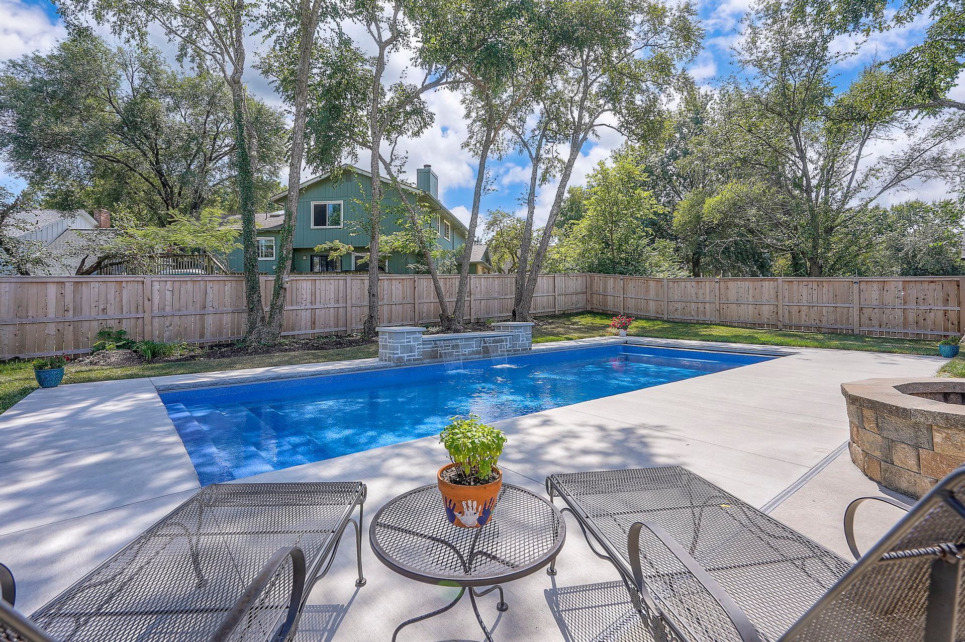 Backyard with a pool, concrete patio, lounge chairs, small table with a potted plant, and a fire pit.