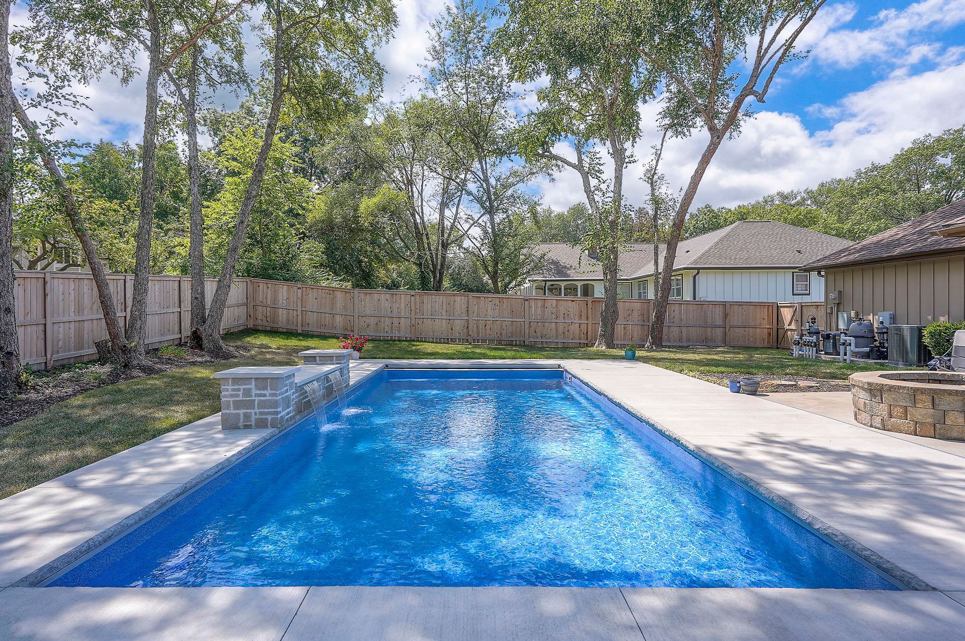 Rectangular pool with blue water in a backyard. Concrete patio, wooden fence, trees, and houses in the background.