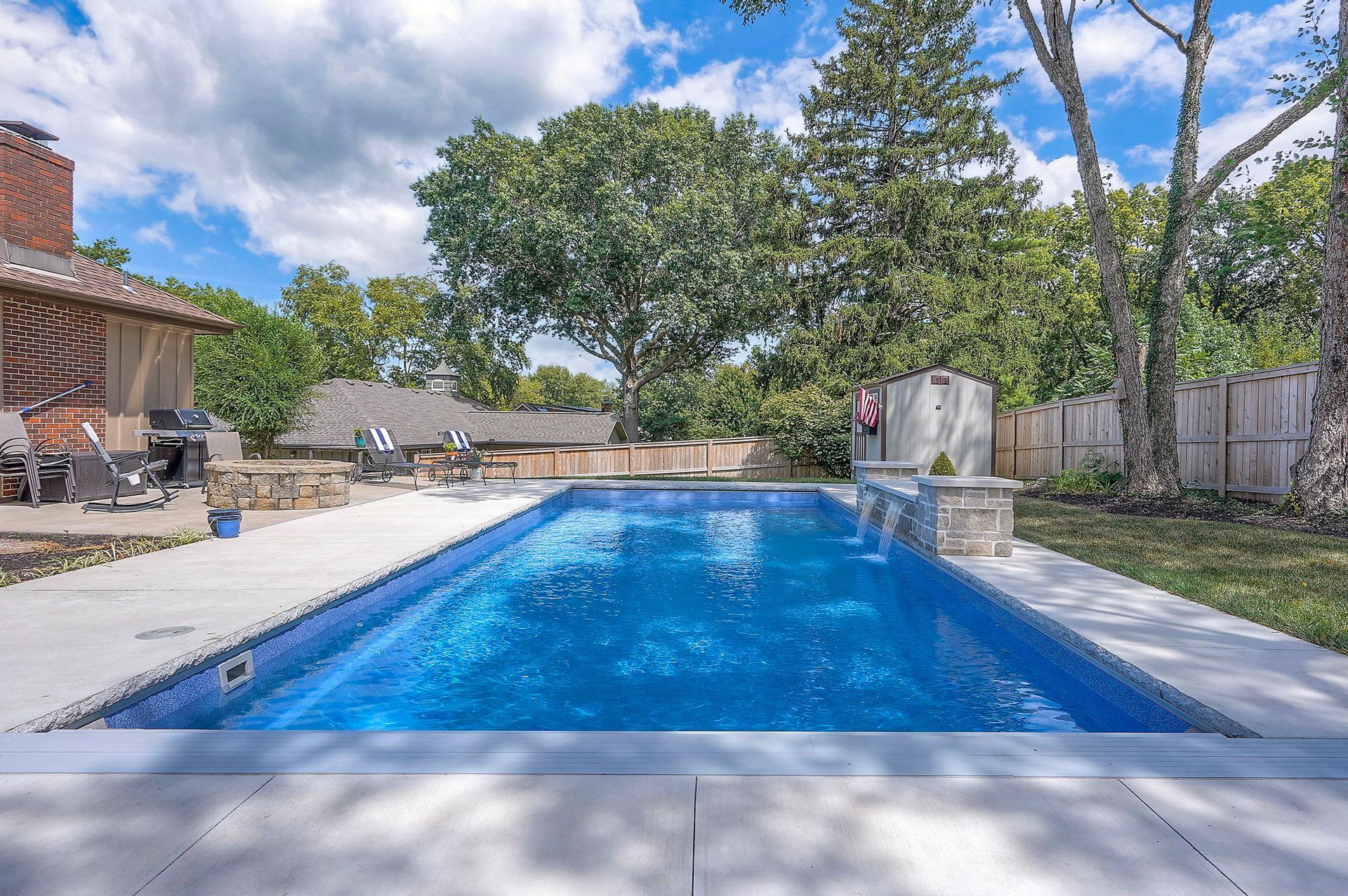 Rectangular swimming pool with blue water, surrounded by concrete patio and greenery under a blue sky.