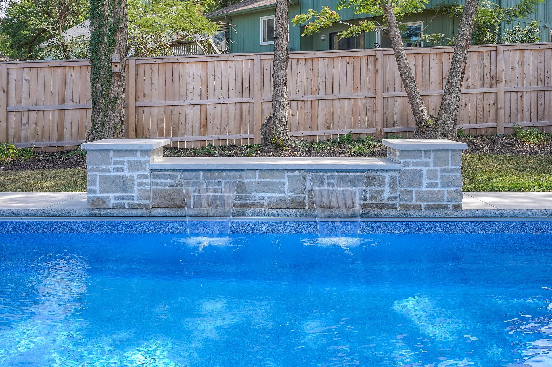 Blue pool with waterfall feature and a wooden fence backdrop.