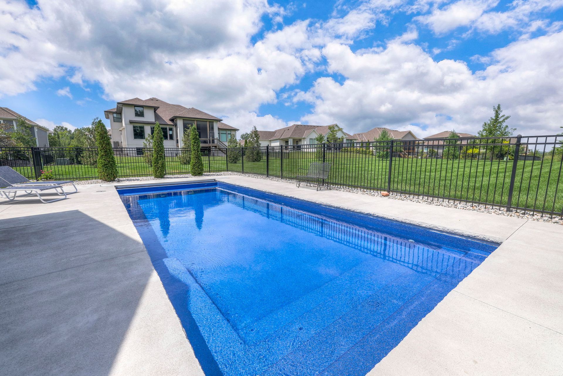 Rectangular blue swimming pool in a concrete patio, with houses and blue sky in background.