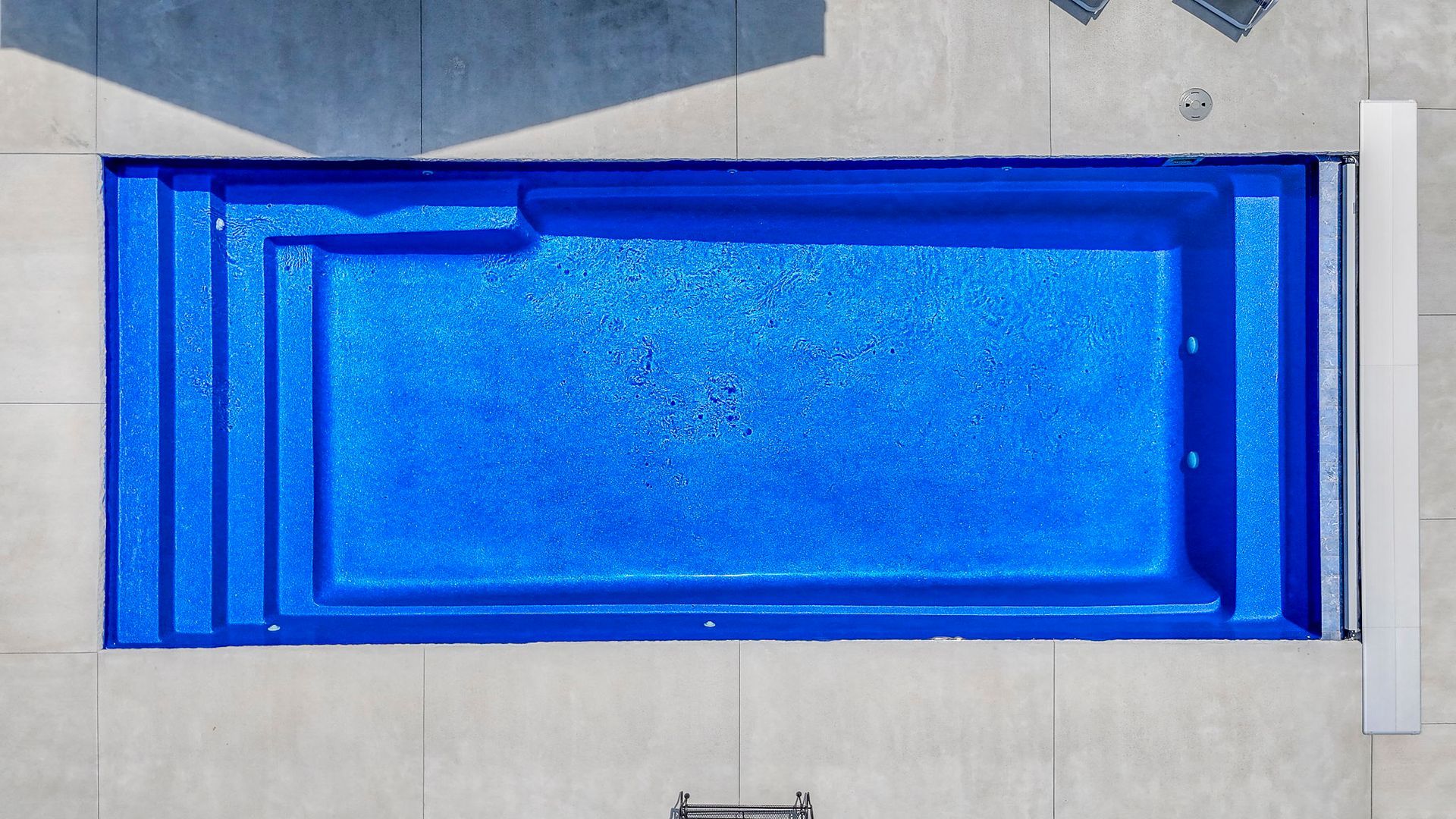 Overhead view of a rectangular, blue-tiled swimming pool with steps, surrounded by concrete.