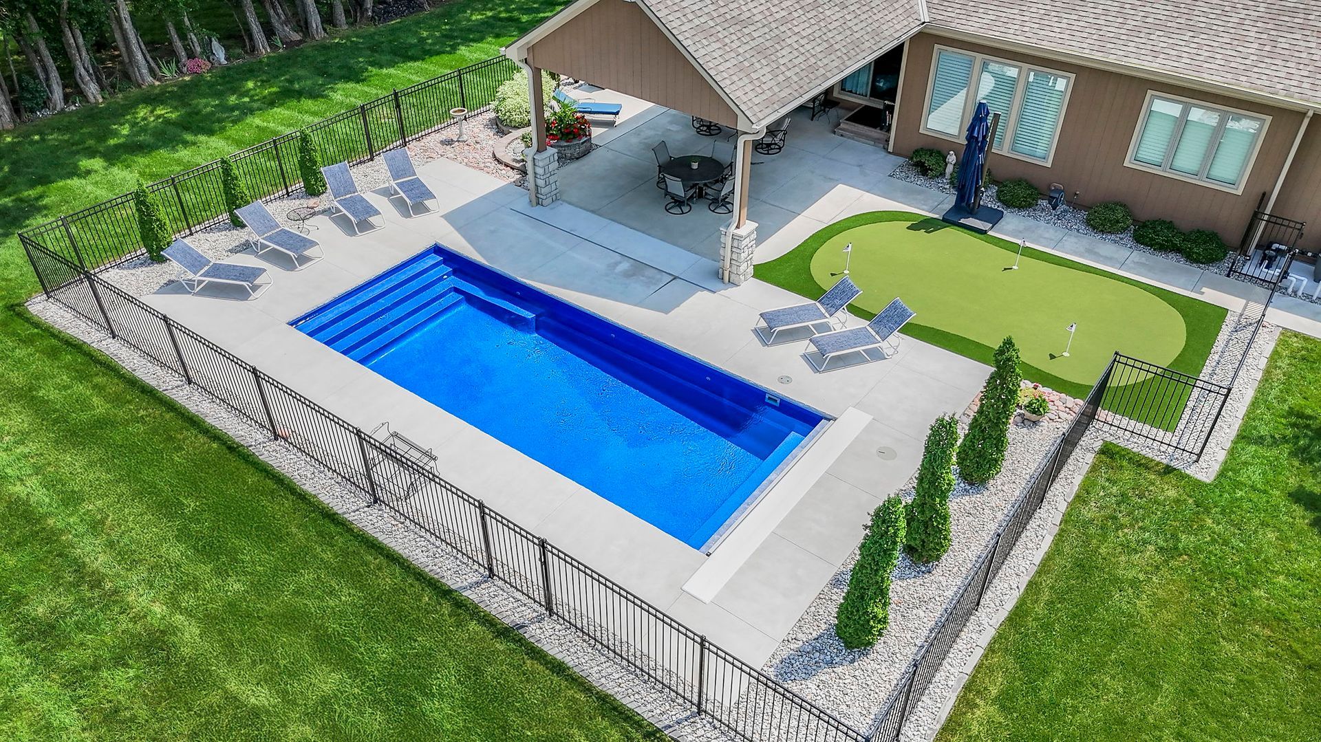 Aerial view of a rectangular blue swimming pool surrounded by concrete, lawn, and a patio, with lounge chairs.