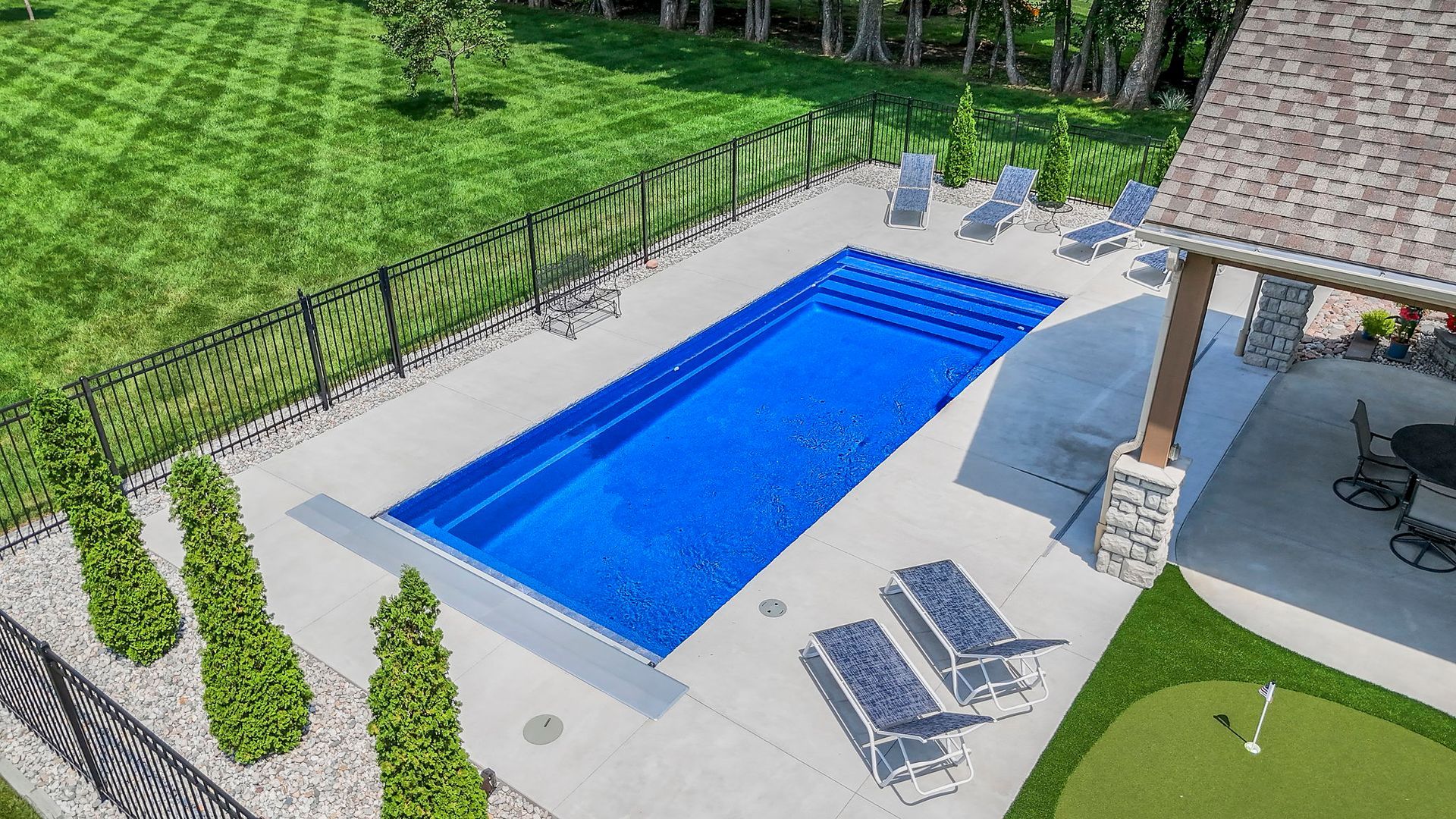 Aerial view of rectangular blue pool with concrete patio, lawn, and bordering black fence.