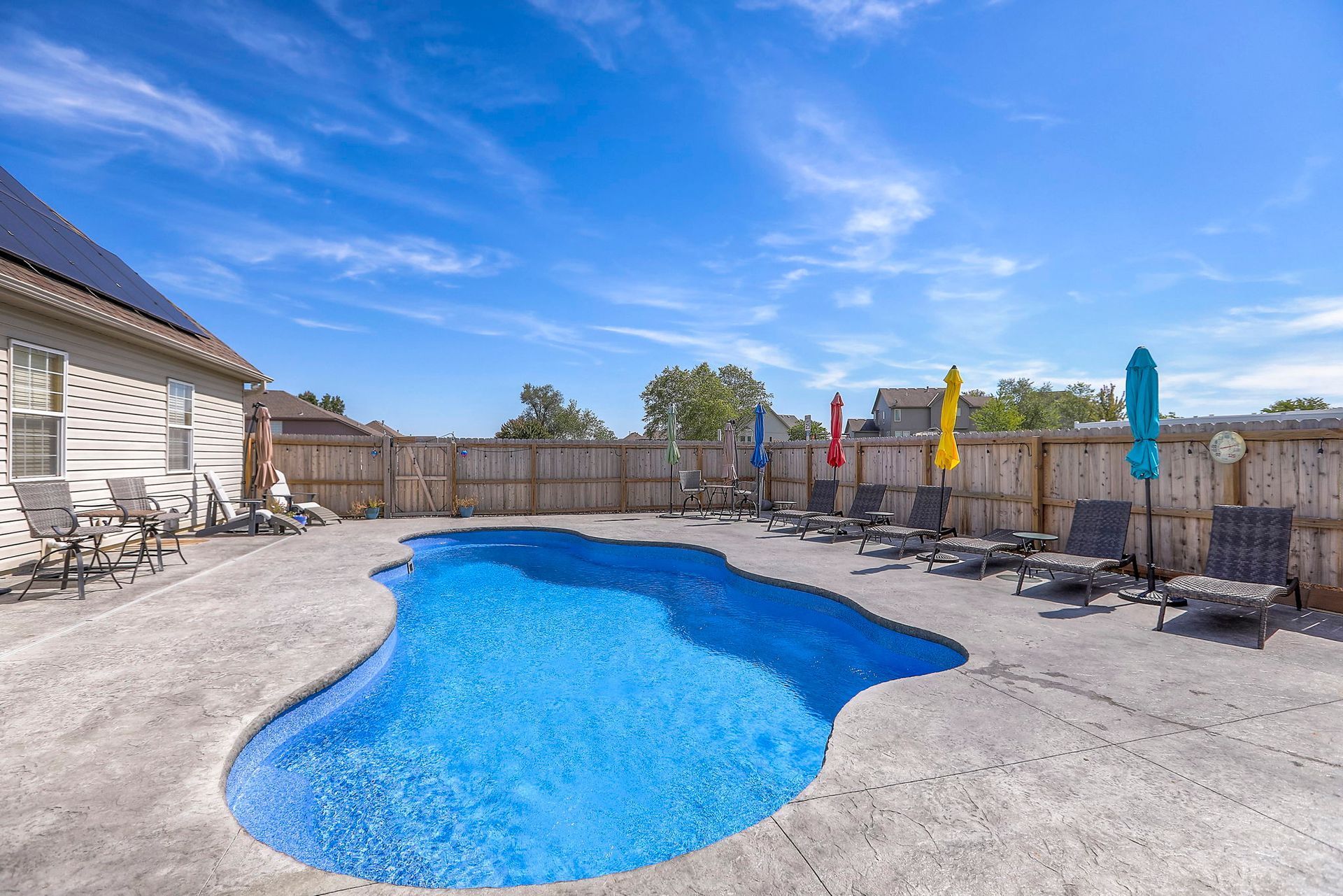 A swimming pool with blue water surrounded by concrete, lounge chairs, and colorful umbrellas under a blue sky.