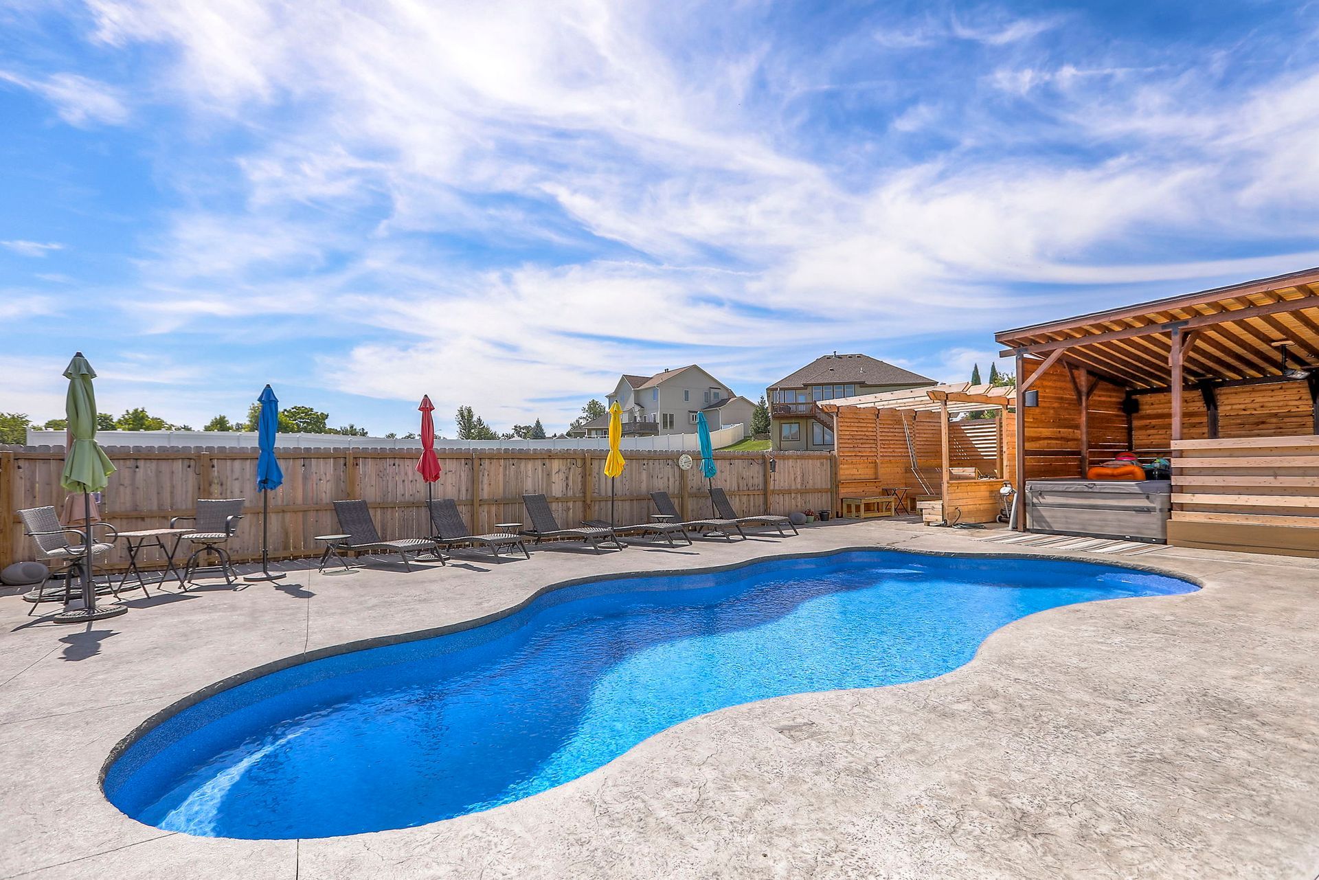 Backyard with blue freeform pool, lounge chairs, colorful umbrellas, and wooden cabana on a sunny day.