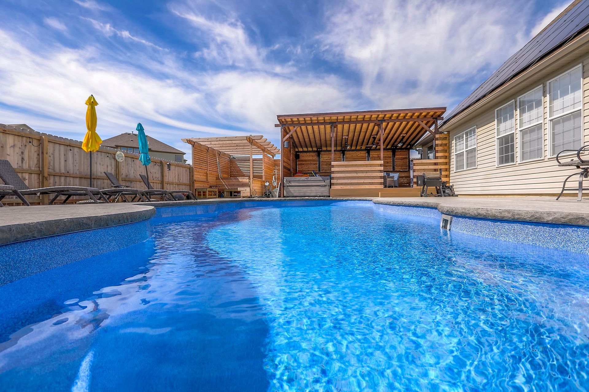 Swimming pool with blue water, wooden cabanas, and a partly cloudy sky.
