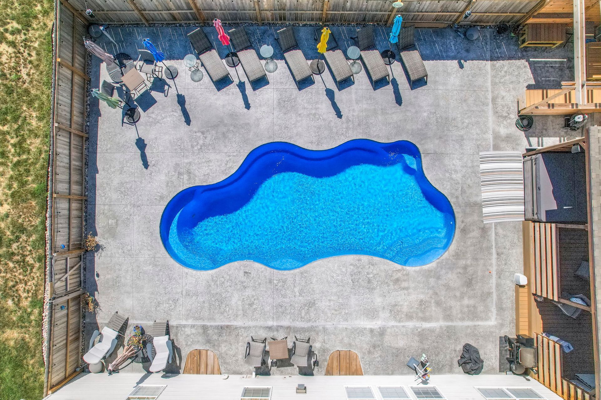 Aerial view of a swimming pool with lounge chairs on a concrete patio, fenced in yard.