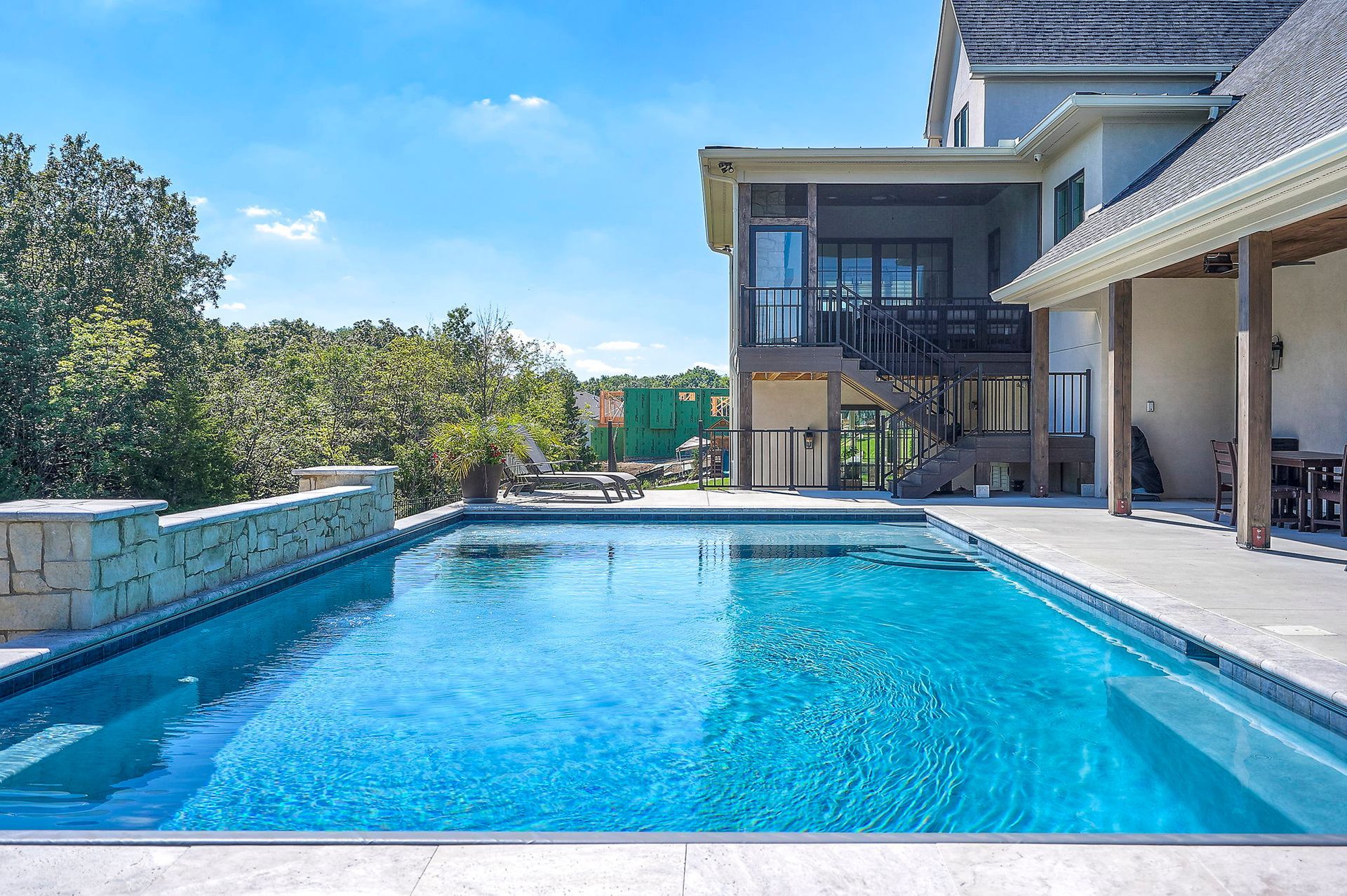 Pool with blue water next to a multi-story house and trees under a partly cloudy sky.