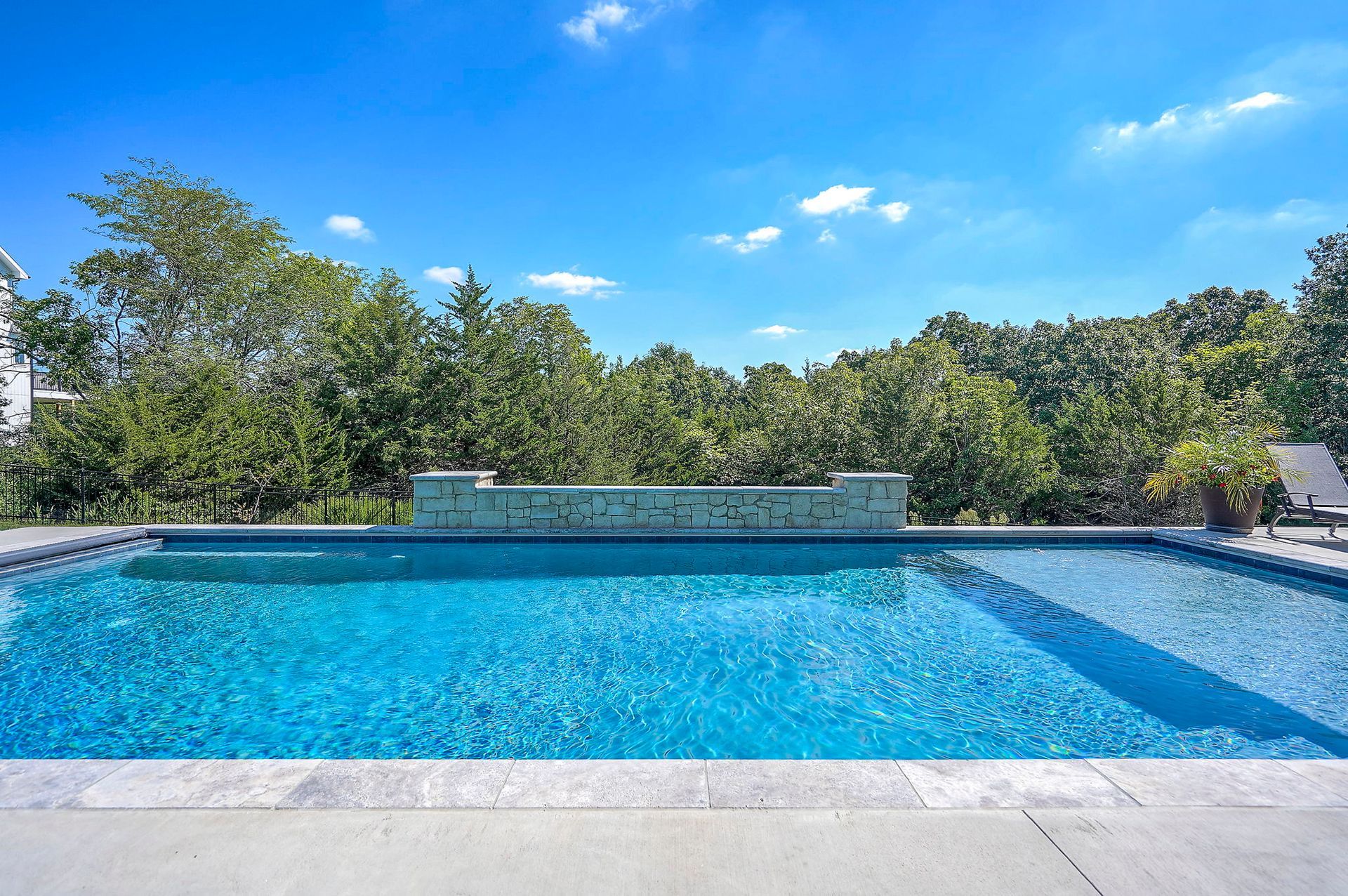 Swimming pool with blue water and stone deck under a bright blue sky, surrounded by trees.