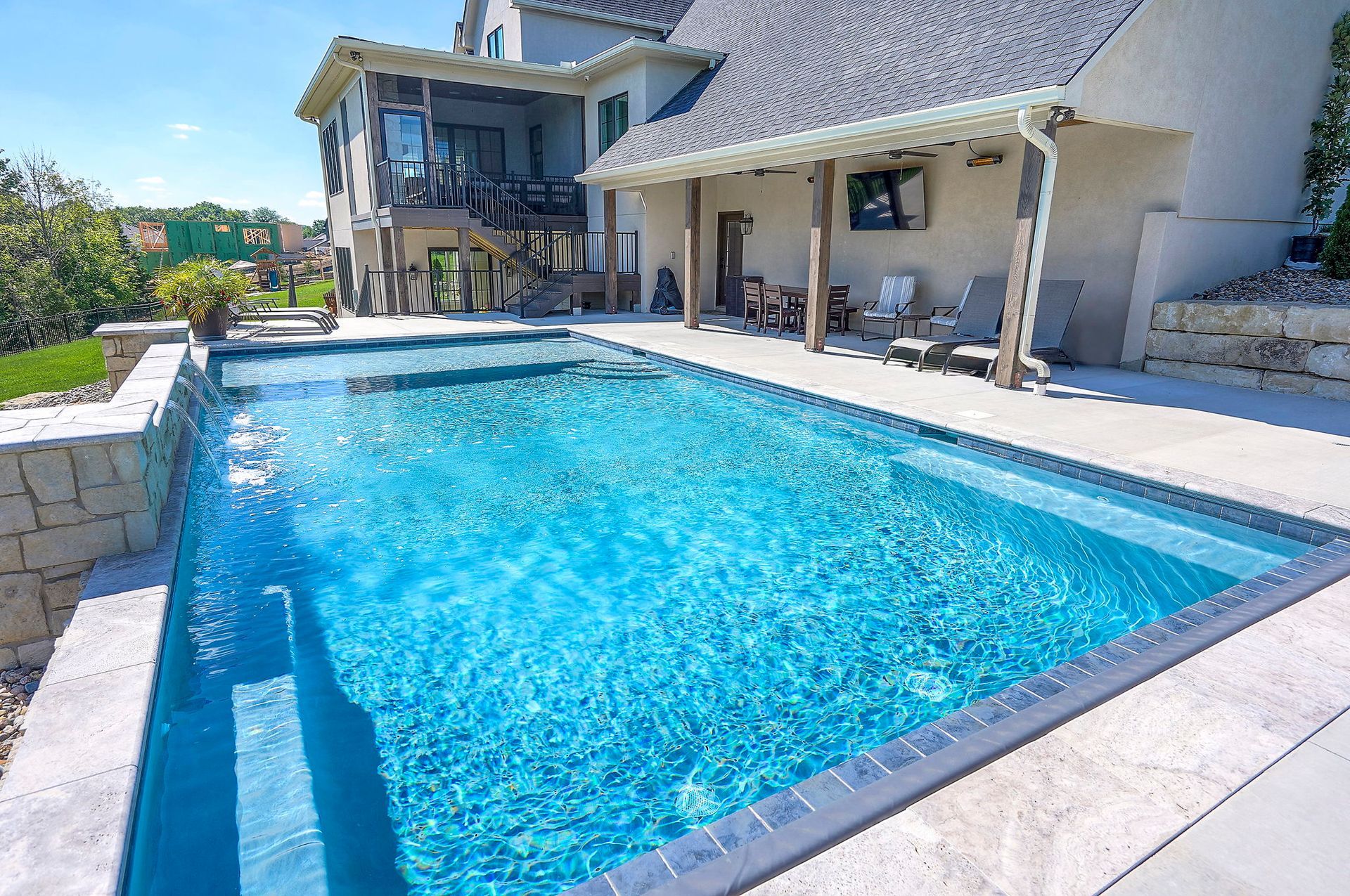 Pool with blue water and steps, beside a light-colored house with a shaded patio.