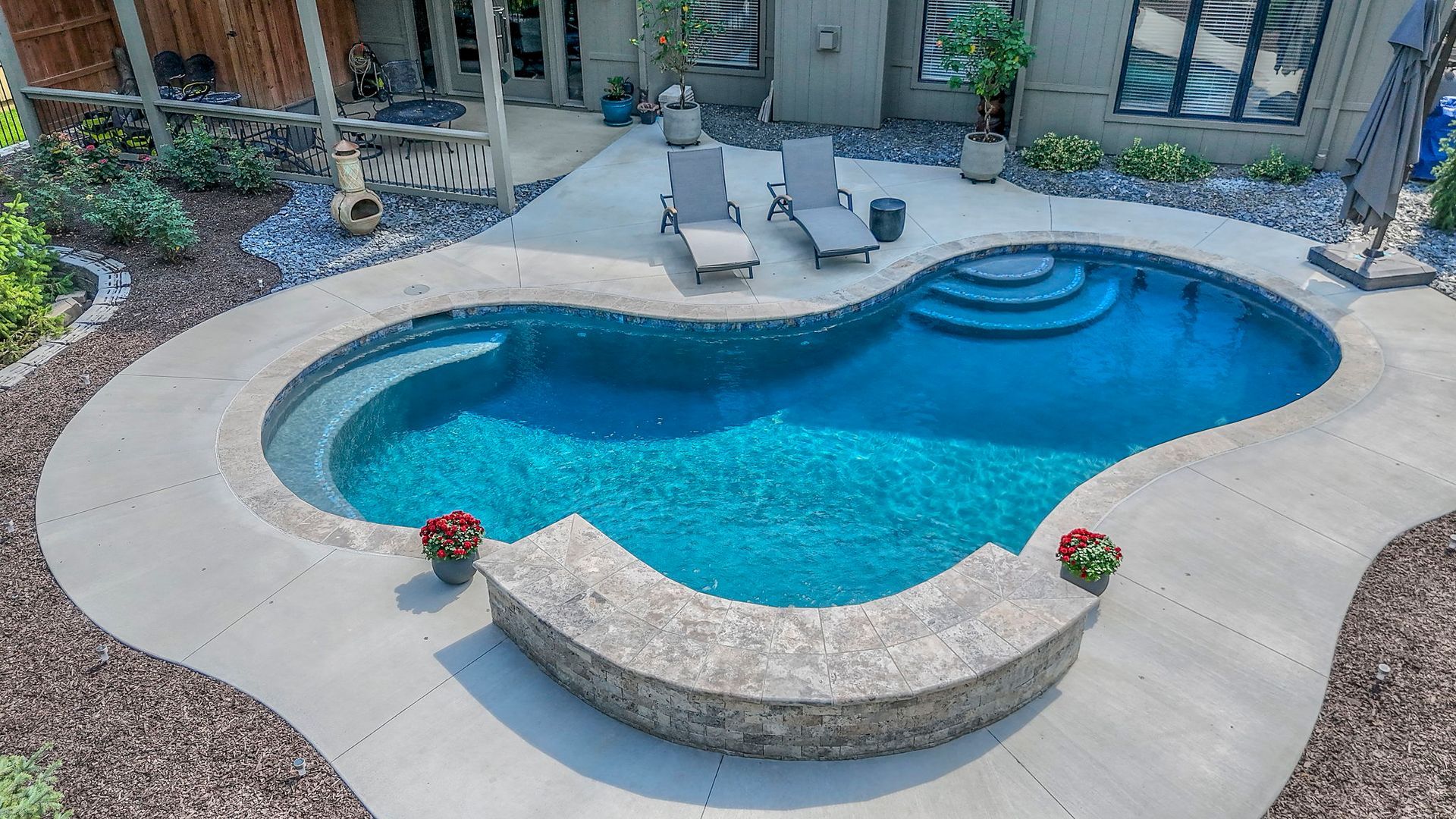 A kidney-shaped swimming pool with a stone wall and concrete patio, lounge chairs, and landscaping.