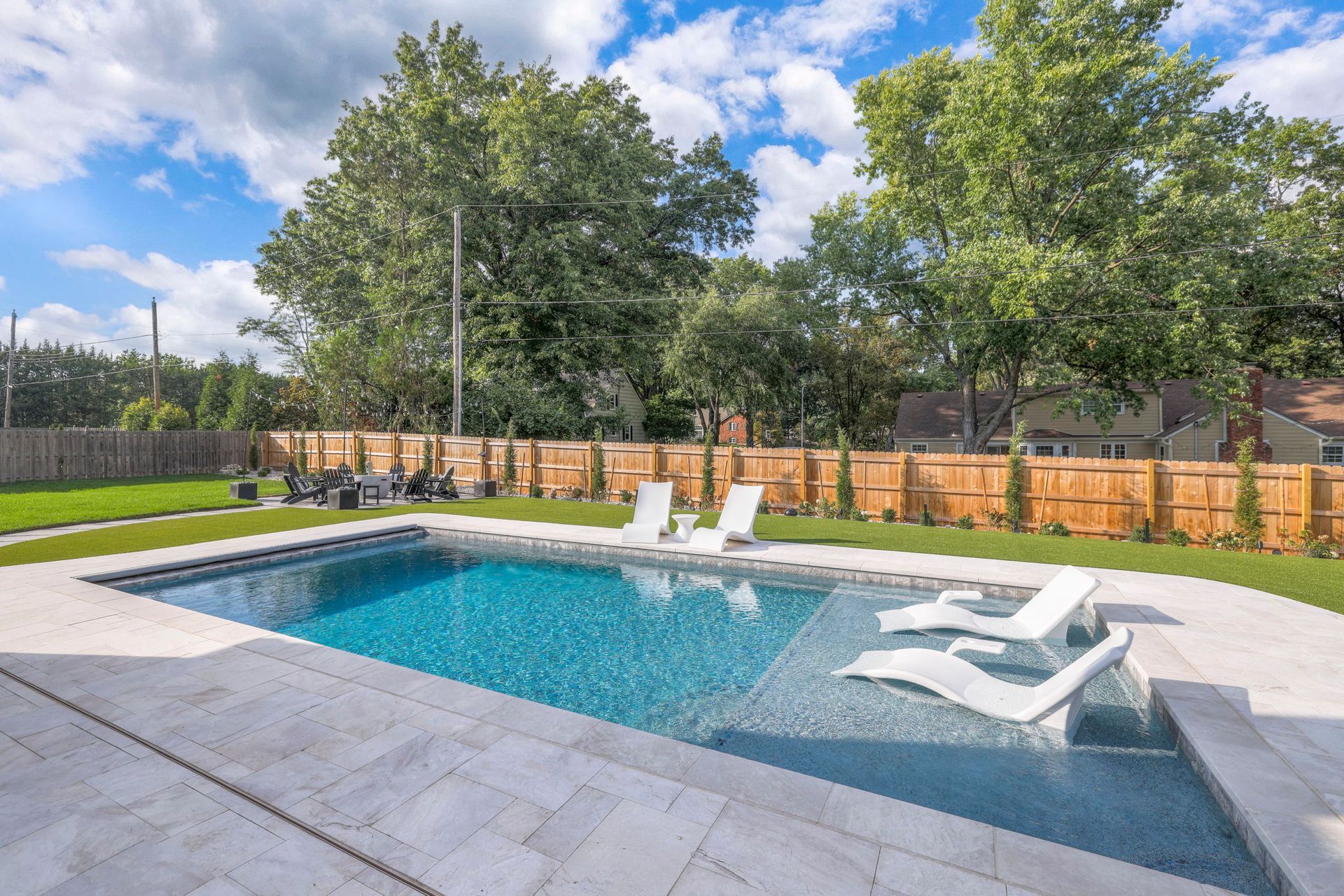 Pool with blue water and white lounge chairs; trees and fence in the background.
