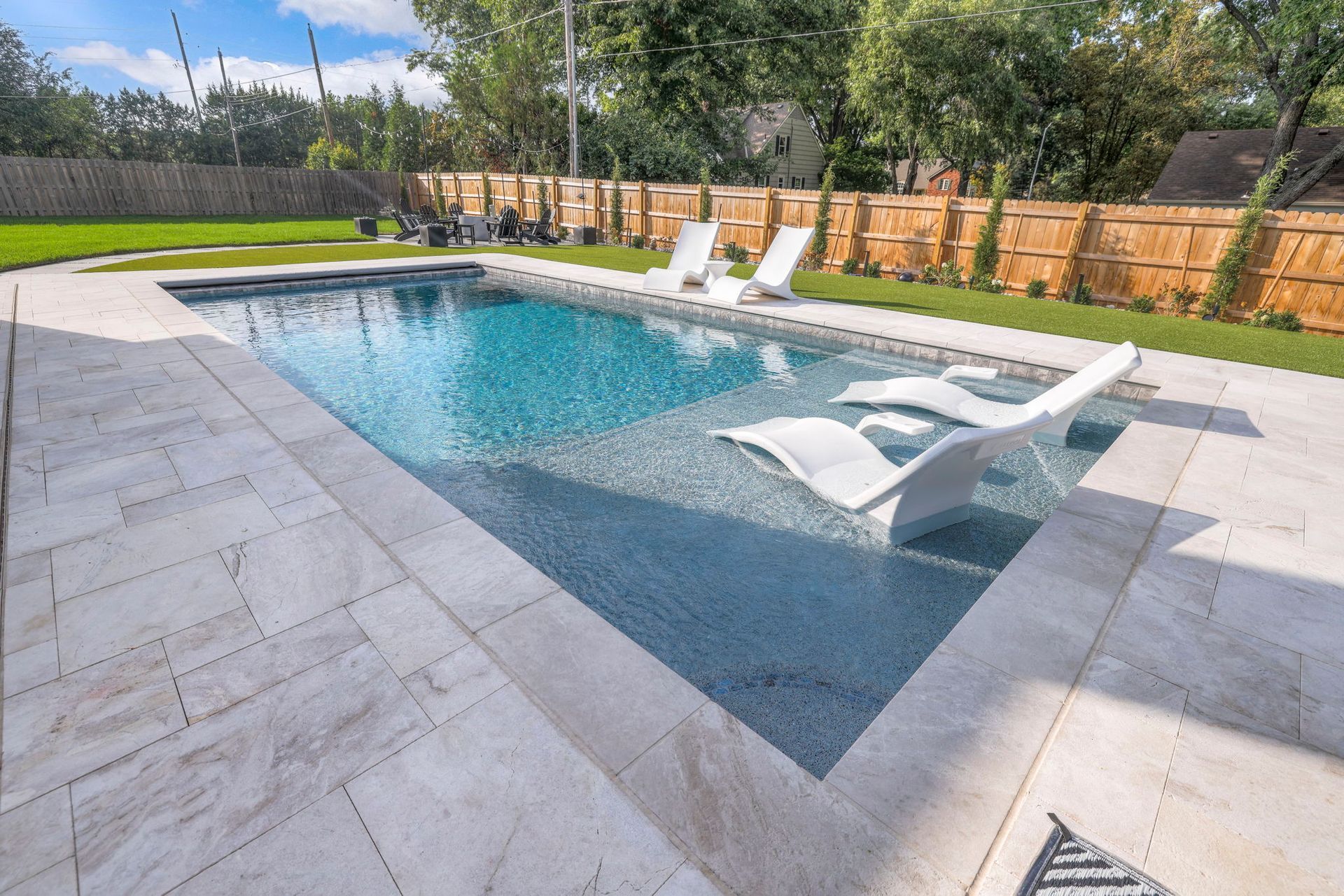 Rectangular pool with white lounge chairs, surrounded by light-colored paving stones and green grass. Bamboo fence in background.