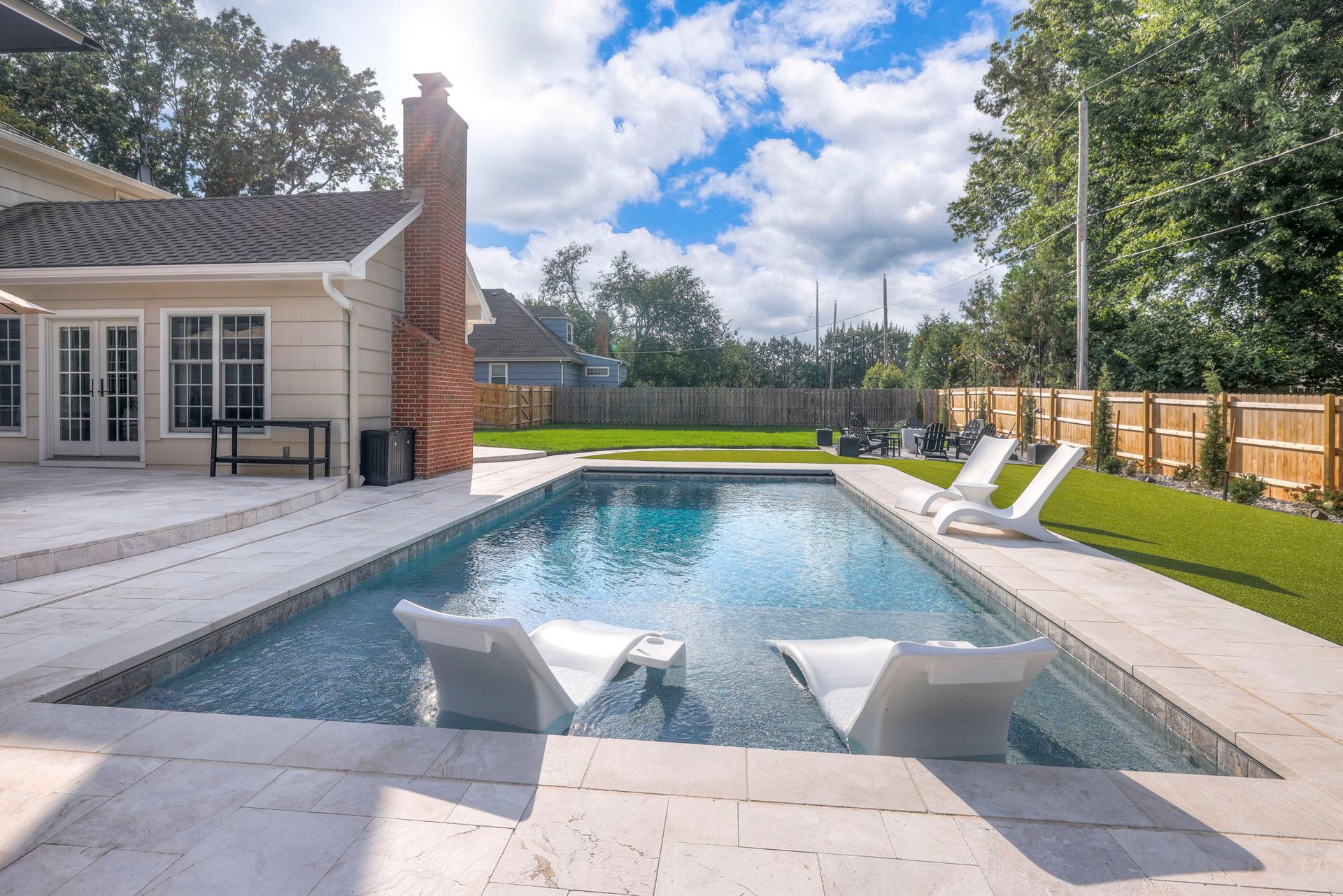 Pool with two white lounge chairs, surrounded by a patio and lawn, under a cloudy sky.