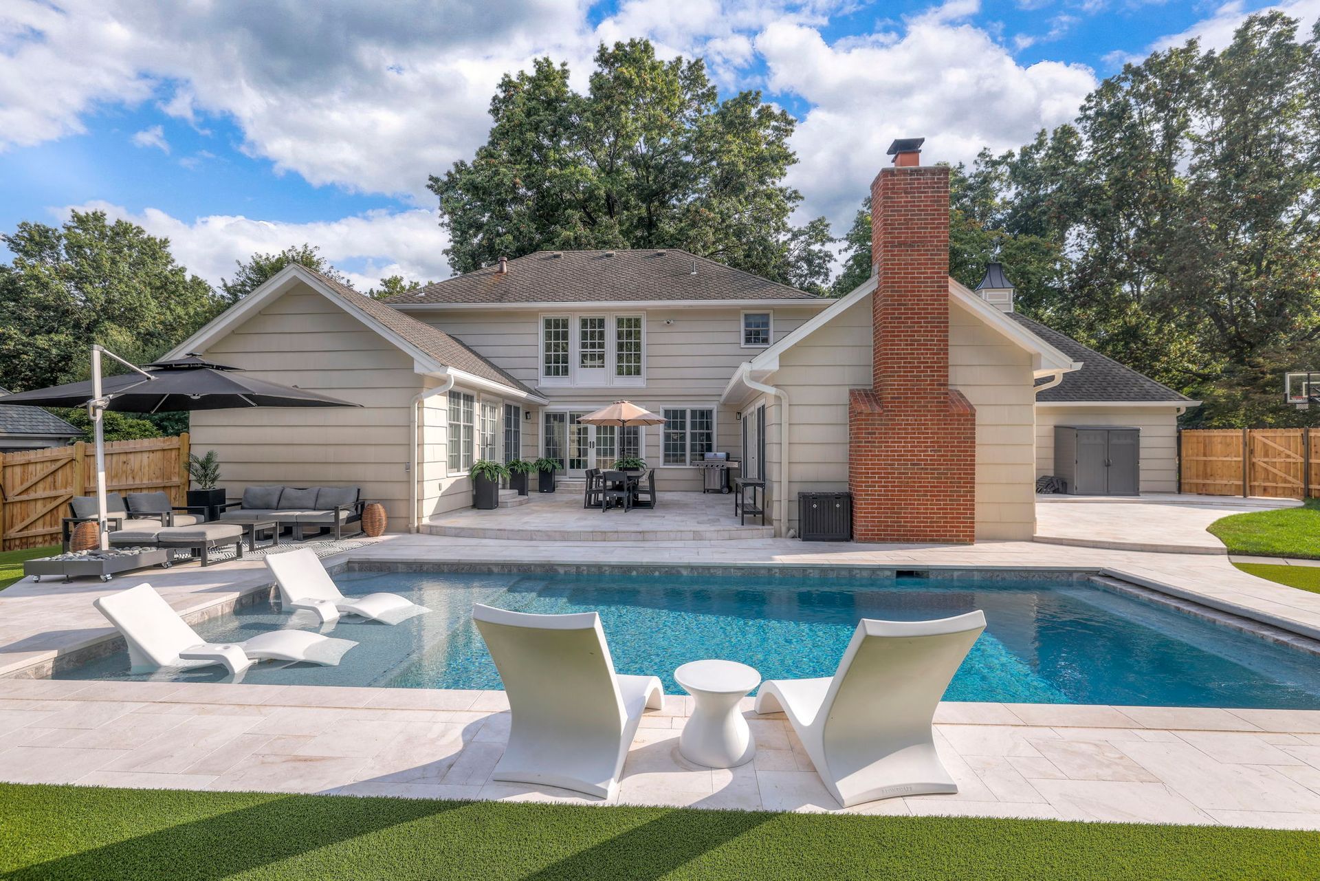 Backyard with pool, patio, and two-story house. White lounge chairs face the pool, blue sky, and green grass.