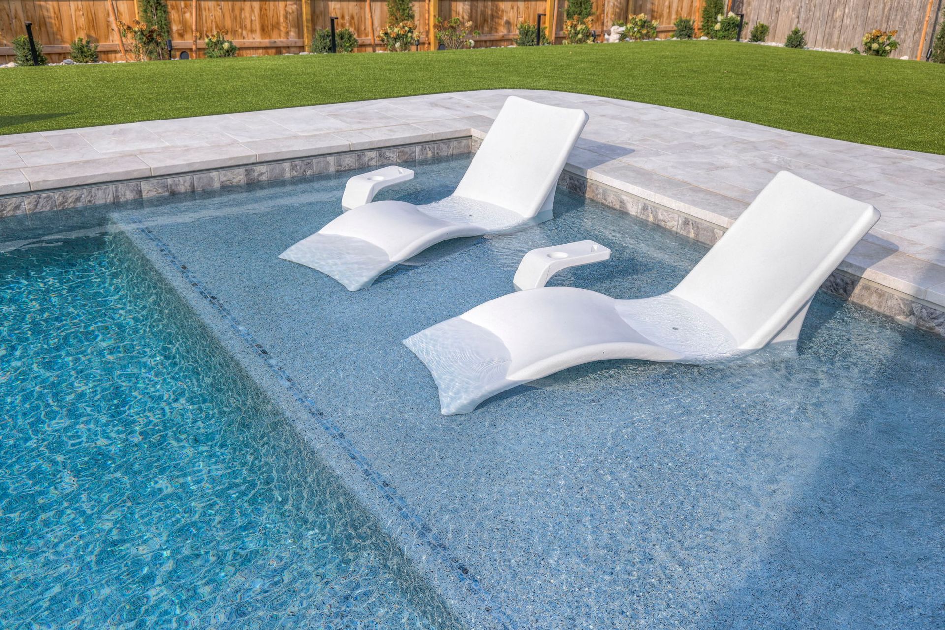 Two white chaise lounge chairs in a shallow pool with clear blue water; lawn and fence in the background.