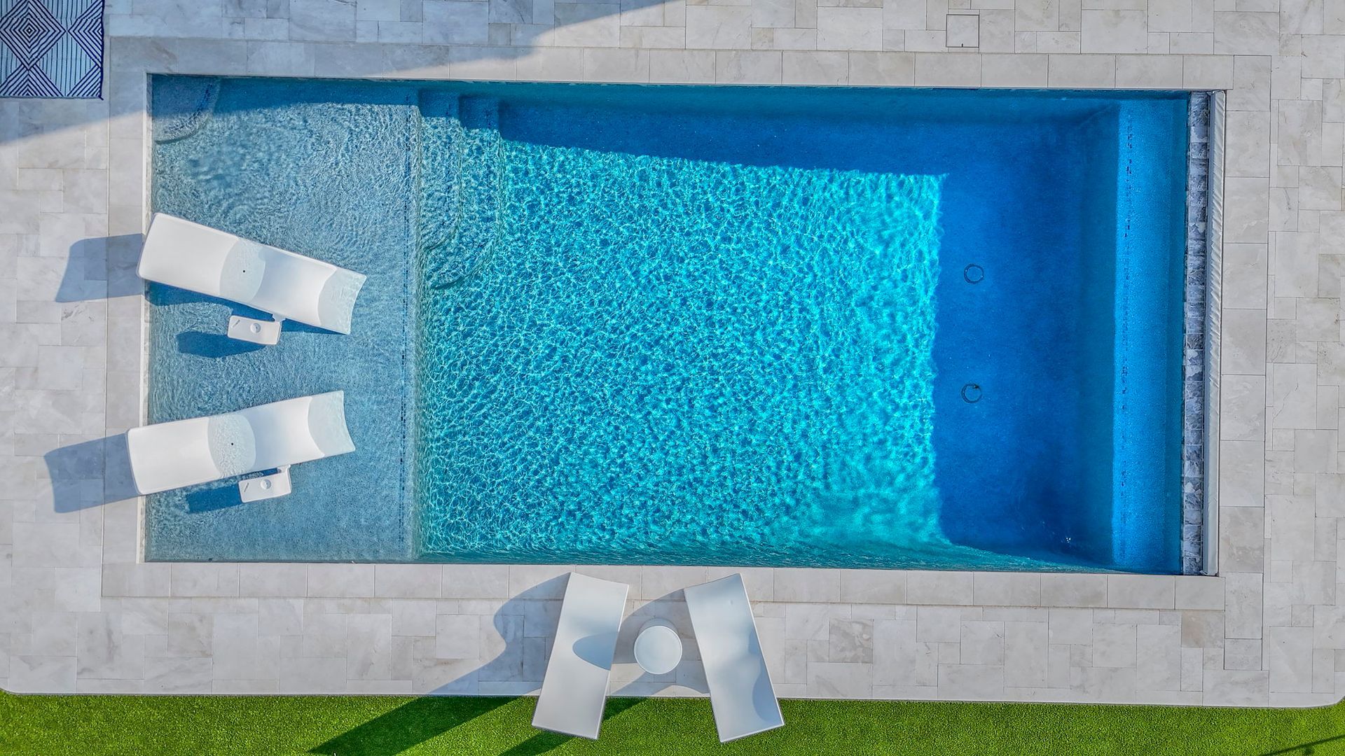 Overhead view of a rectangular blue swimming pool with two white lounge chairs and a matching table on a patio.