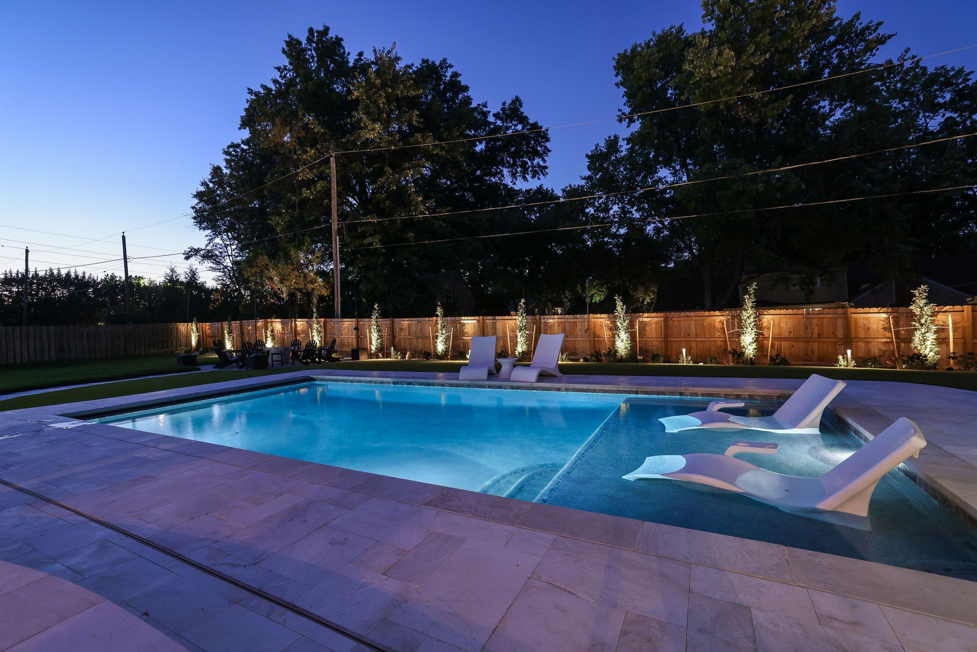 Nighttime view of a pool with in-water lounge chairs; string lights and trees in the background.