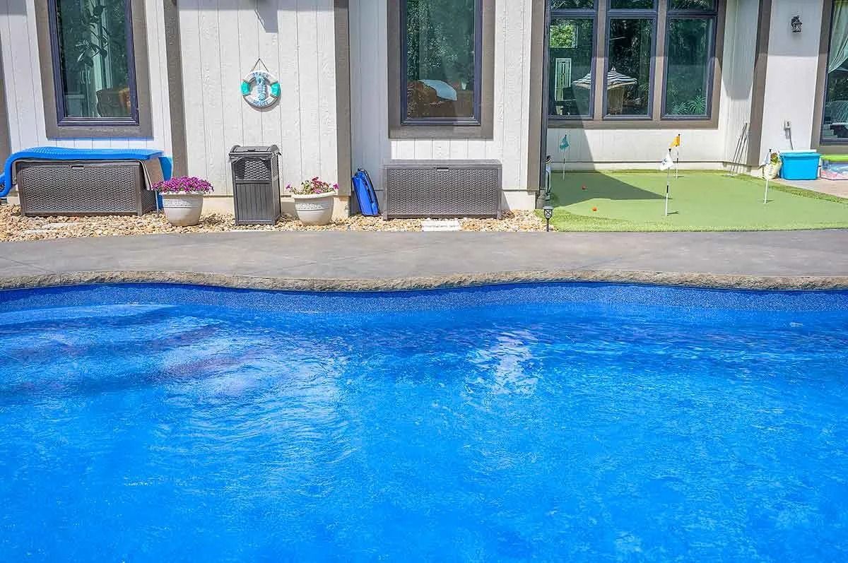 Blue pool in front of a white house with dark window frames. Artificial grass and a putting green are visible.