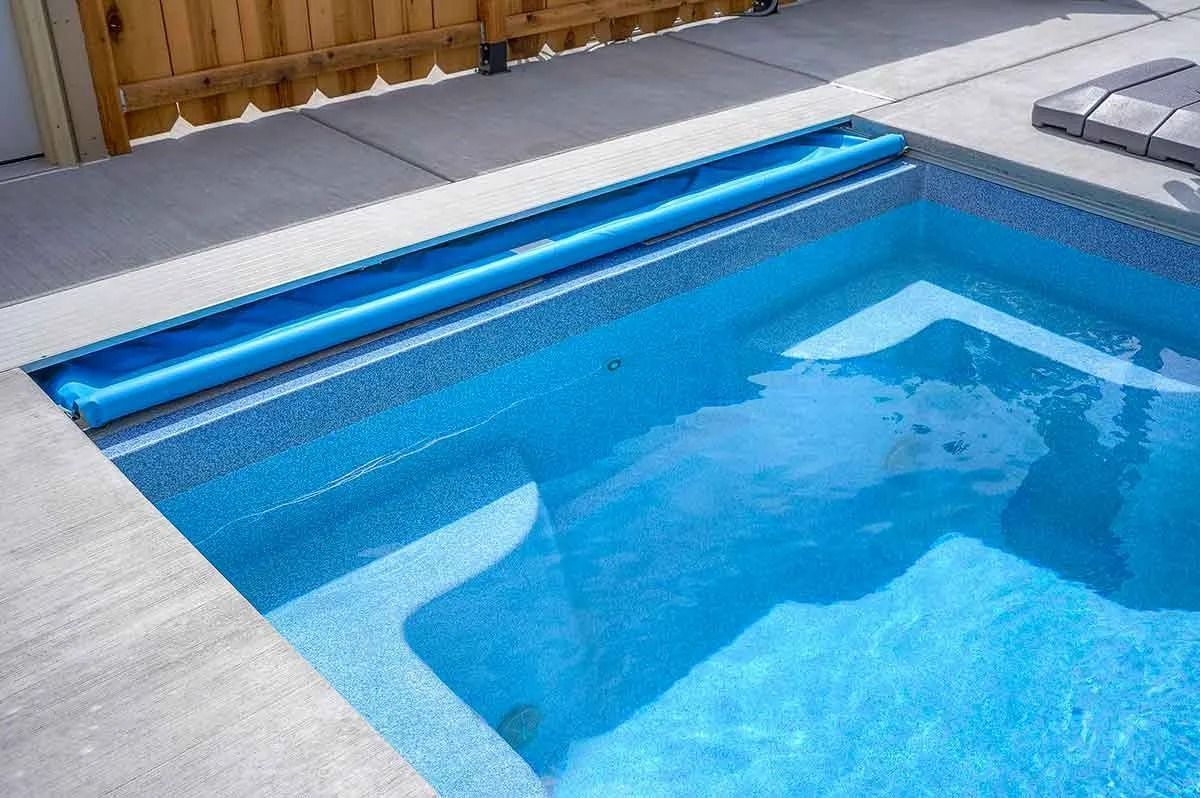 Blue pool with water and steps, blue cover rolled up near edge, light gray concrete border.