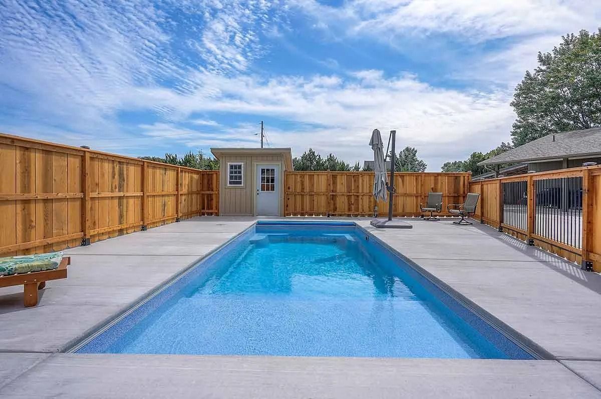 Rectangular blue pool surrounded by a wooden fence and concrete. A small wooden shed is visible. Sunny, daytime.