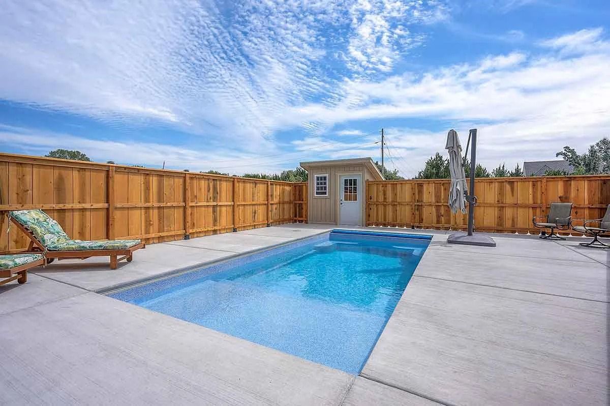 Rectangular backyard pool with a shed, wooden fence, and lounge chairs under a blue sky.