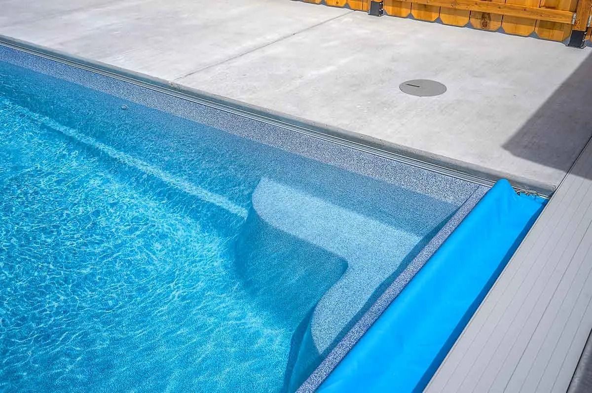 Swimming pool with blue water and steps, blue cover, concrete patio, and wood fence.