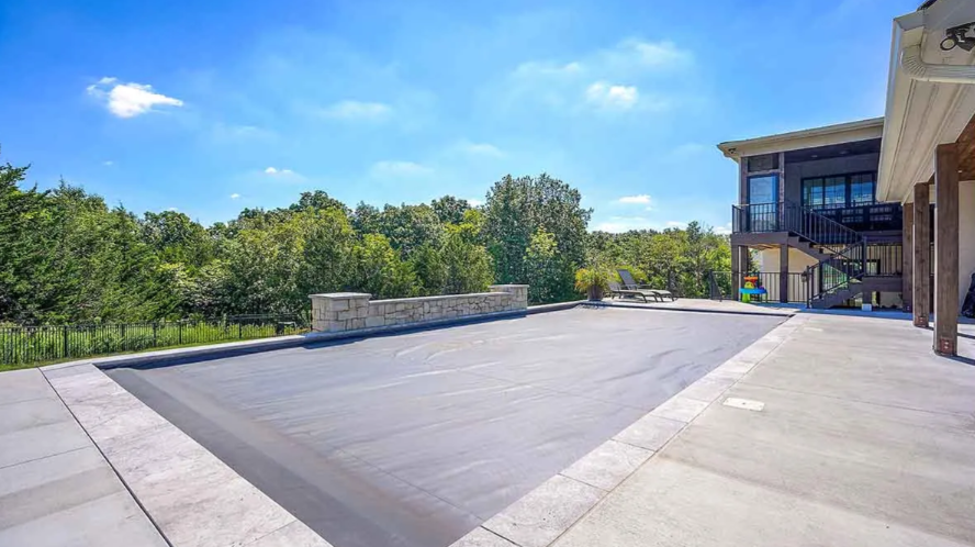 Pool covered in gray material next to a house with a green landscape and blue sky.