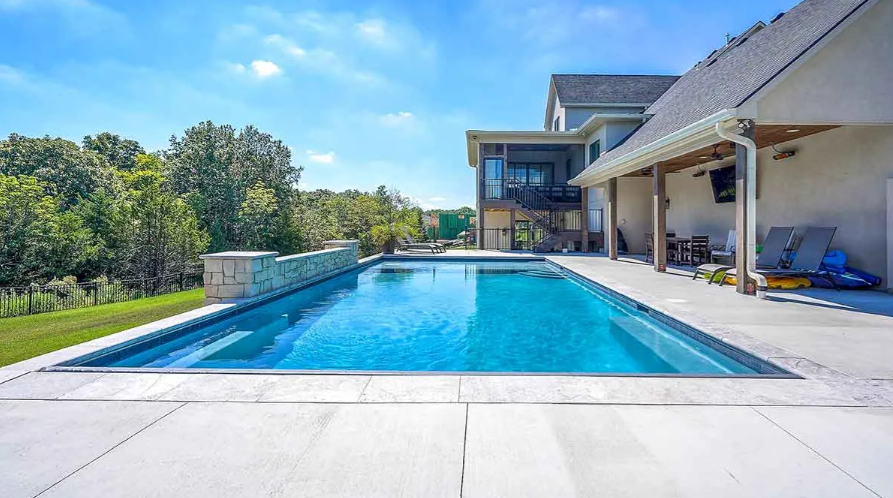 Pool next to a house with a deck and porch. Blue sky, water, and green trees.