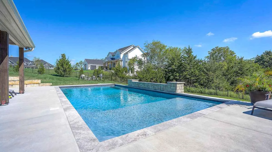 Rectangular outdoor pool with blue water on a sunny day. A house and trees are in the background.