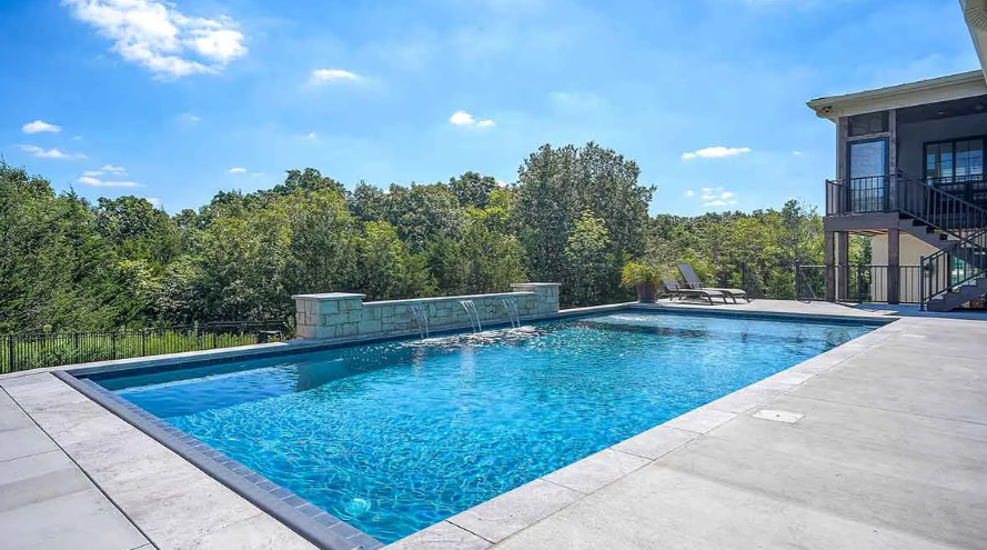 Swimming pool with clear blue water and stone wall, backed by green trees and blue sky.