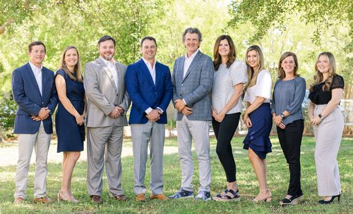 Eight people in business attire standing together outdoors in a sunny park-like setting.
