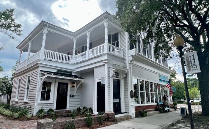 White two-story corner shop with wraparound porch and balcony under trees on a city street