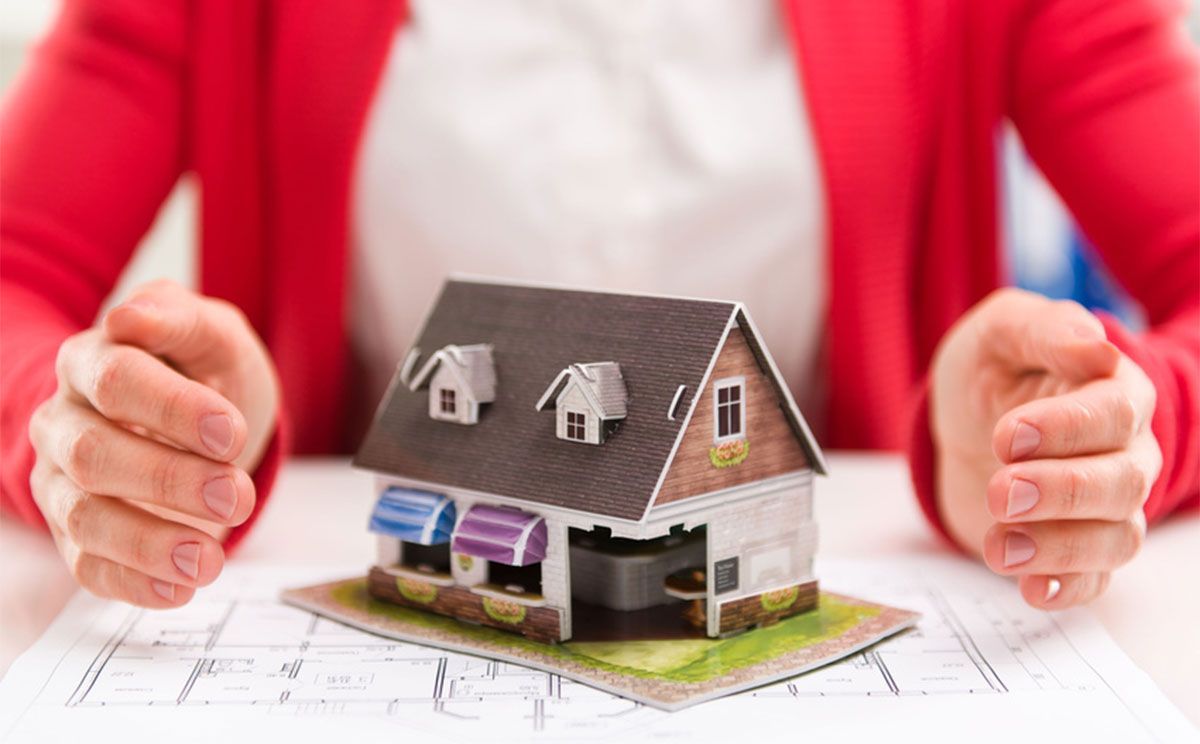 Hands framing a small model house on a blueprint, with a red jacket in the background