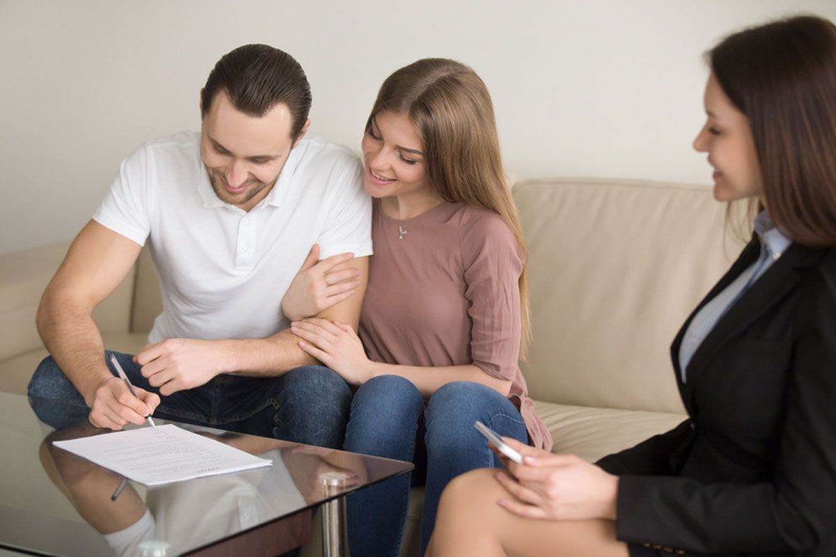 Three people in a meeting; one man signs papers while a woman and advisor watch on a sofa.