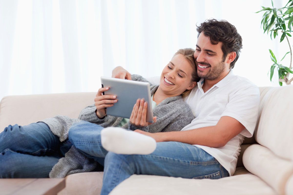Couple cuddling on a white couch and looking at a tablet in a bright living room