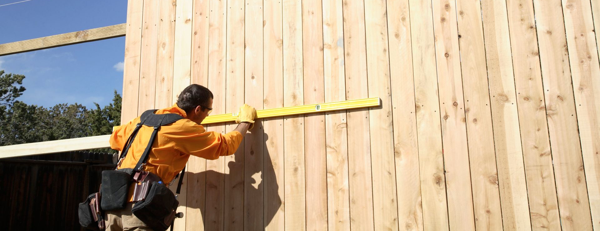 Worker in orange jacket measuring a wooden wall with a yellow tape measure outdoors