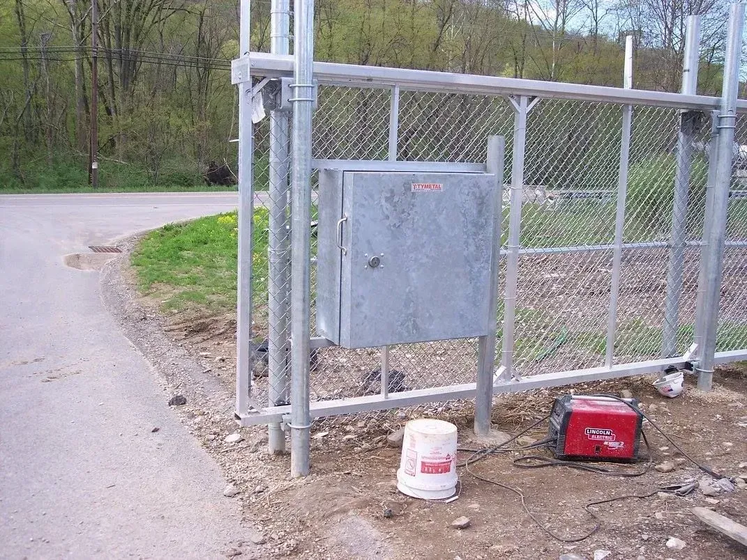 A metal box on a fence post with a gate, by a road, and a white bucket.