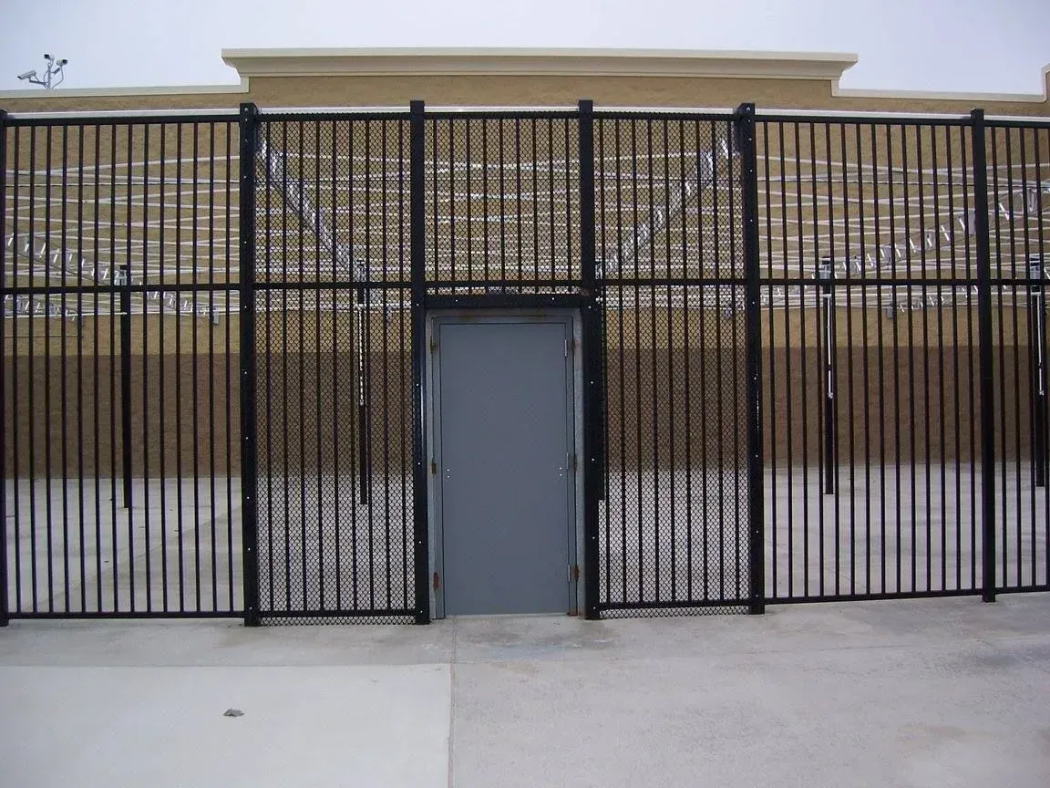 Black security fence with door, topped with razor wire, against a beige wall; exterior shot.