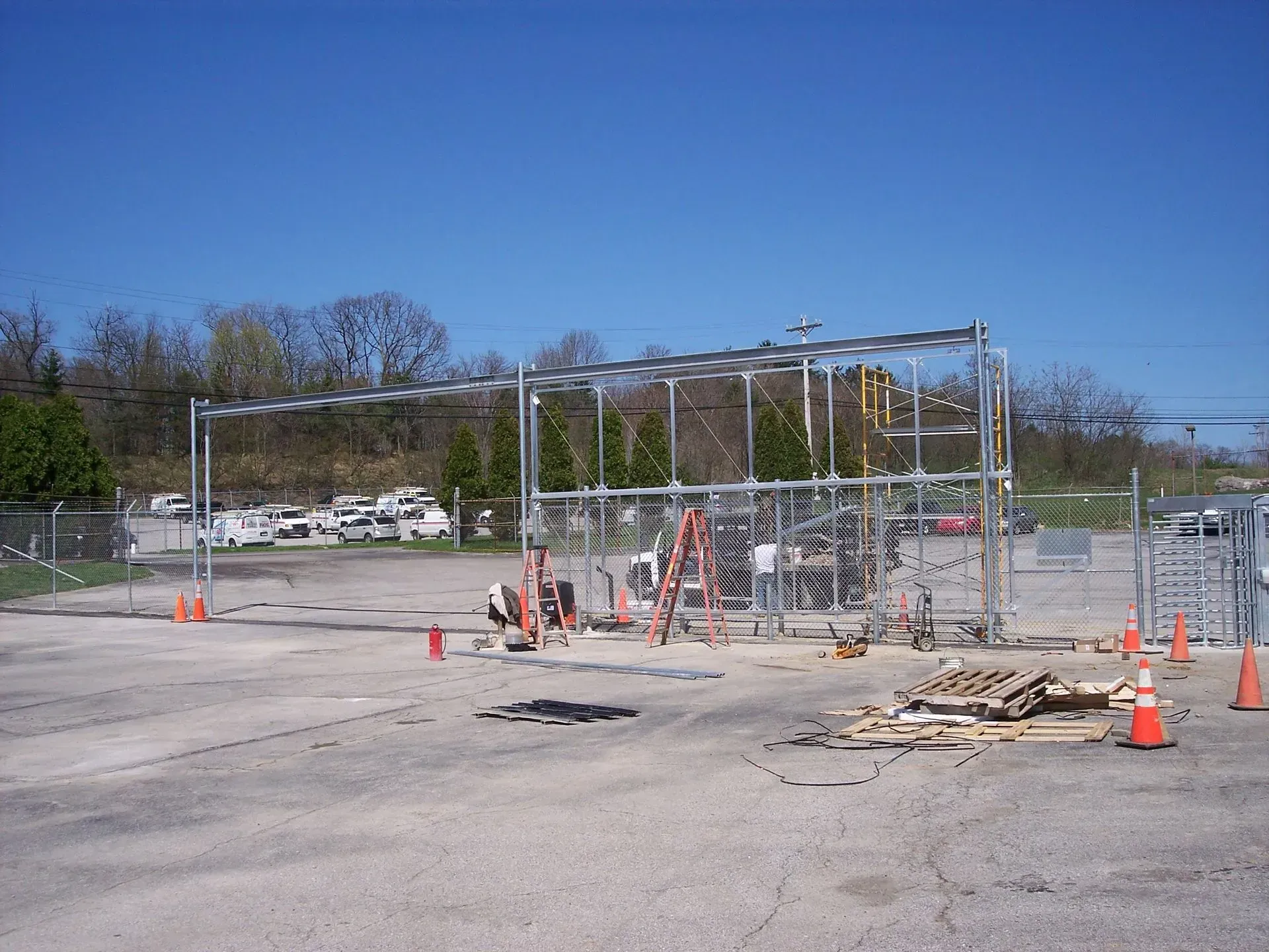 Construction site with metal framework, fence, and equipment. Workers are present on asphalt under a blue sky.
