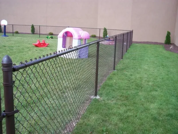 Black chain-link fence encloses a grassy area with a playhouse and toys; set against a beige wall.